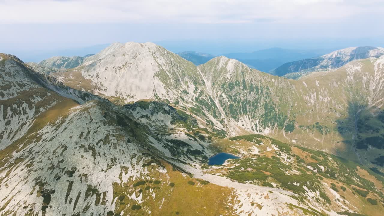 Blue glacial lake in the sunlight, nested between green pastures and mountain rocks, surrounded by tall mountain ranges, aerial tracking shot