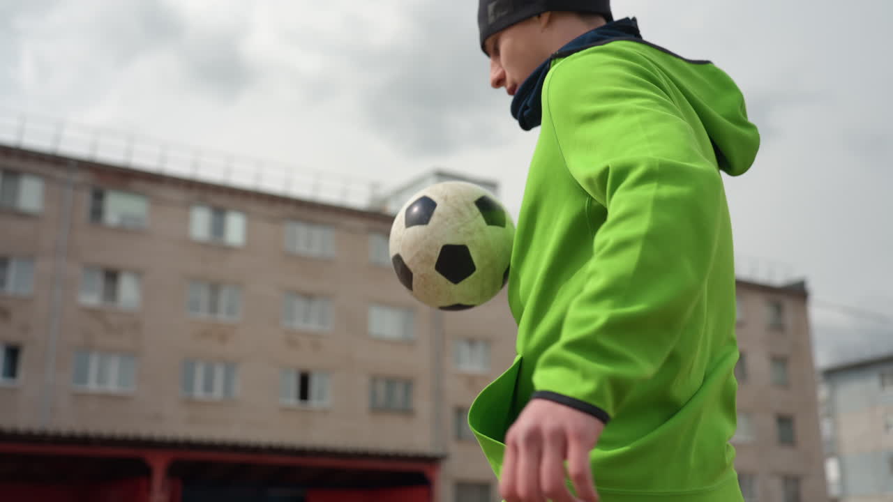 Крупный план: кавказский фристайлер отрабатывает трюки с футбольным мячом возле торса и колена, в зеленой толстовке и серых спортивных штанах, на фоне городской квартиры, игривые техничные движения и уверенный ритм