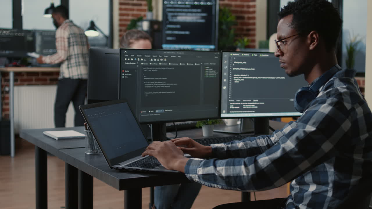 Portrait of tired programer rubbing eyes while typing on laptop sitting at desk with multiple screens parsing code