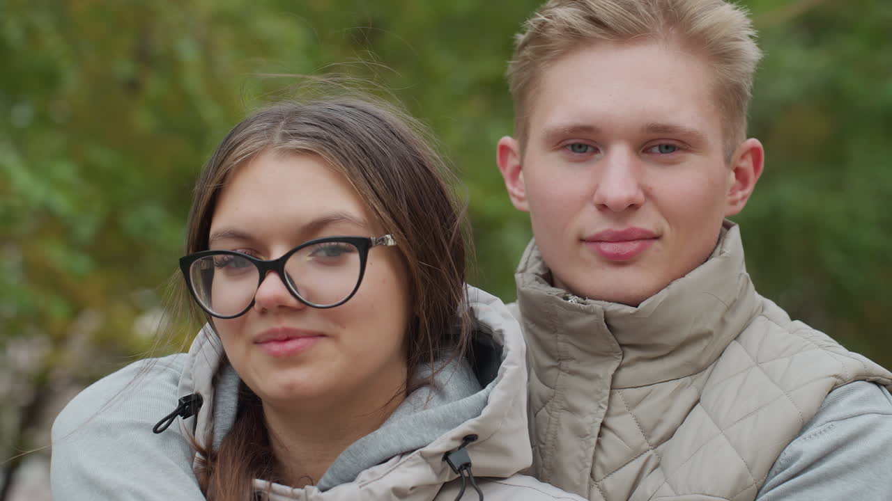 Young man places hand across wife shoulder as they share quiet moment outdoors with eyes closed, wearing matching jackets while tree branches sway in gentle breeze in soft natural background
