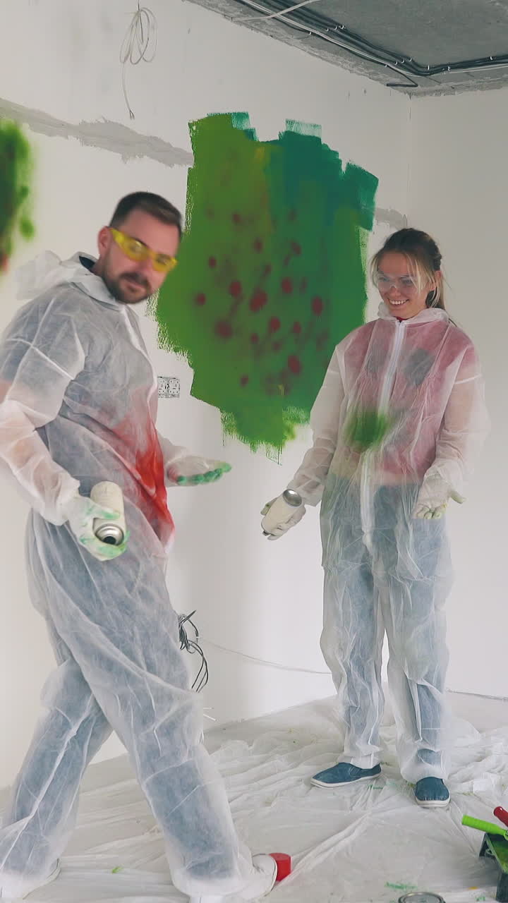 funny young man and woman in white workwear with goggles paint each other with sprays near wall in empty repairing room