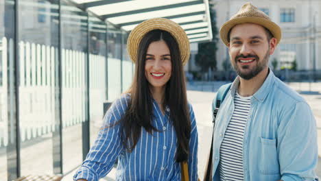 portrait of caucasian young happy couple of travellers in hats with suitcases standing at bus stop and smiling to camera cheerfully