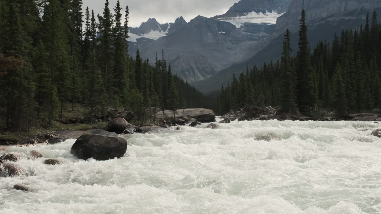 A rushing river, fueled by melting snow, carves through a dramatic mountain valley in the Canadian Rockies