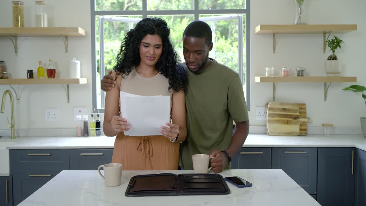 African American man entering kitchen with mugs offering mug, Indian woman reading papers to review