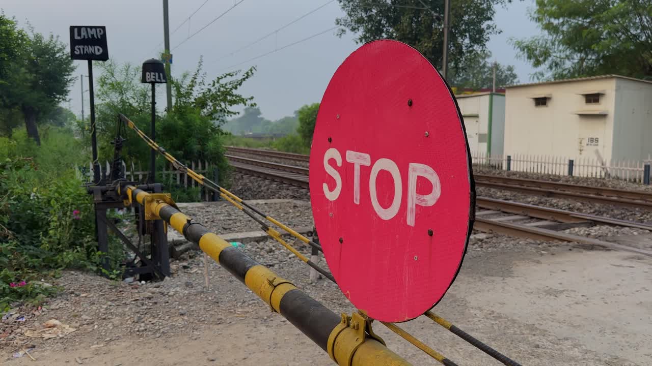 Camera circles around a red stop sign at a railway crossing, showing striped barrier poles, gravel ground, metal tracks, green vegetation, distant buildings, and soft daylight shaping the open scene