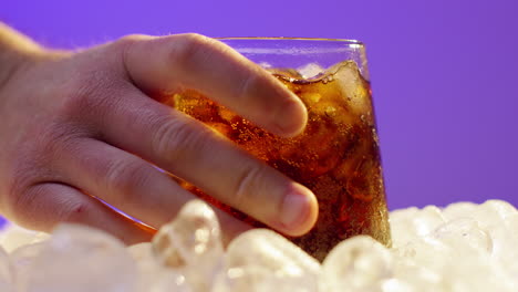 Close-Up-Of-Person-Picking-Up-Chilled-Cold-Drink-In-Glass-On-Ice-Cubes-Against-Purple-Background