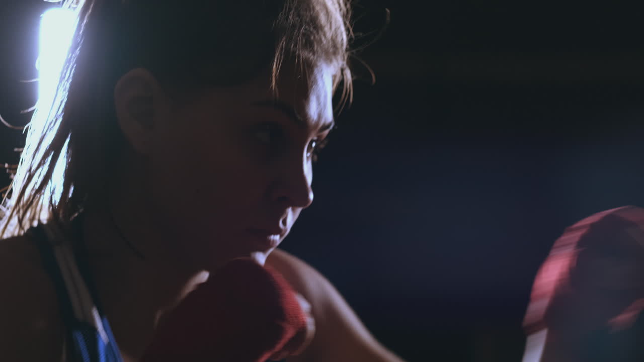 A beautiful woman boxer trains in a dark gym and works out punches in slow motion. side view. Steadicam shot