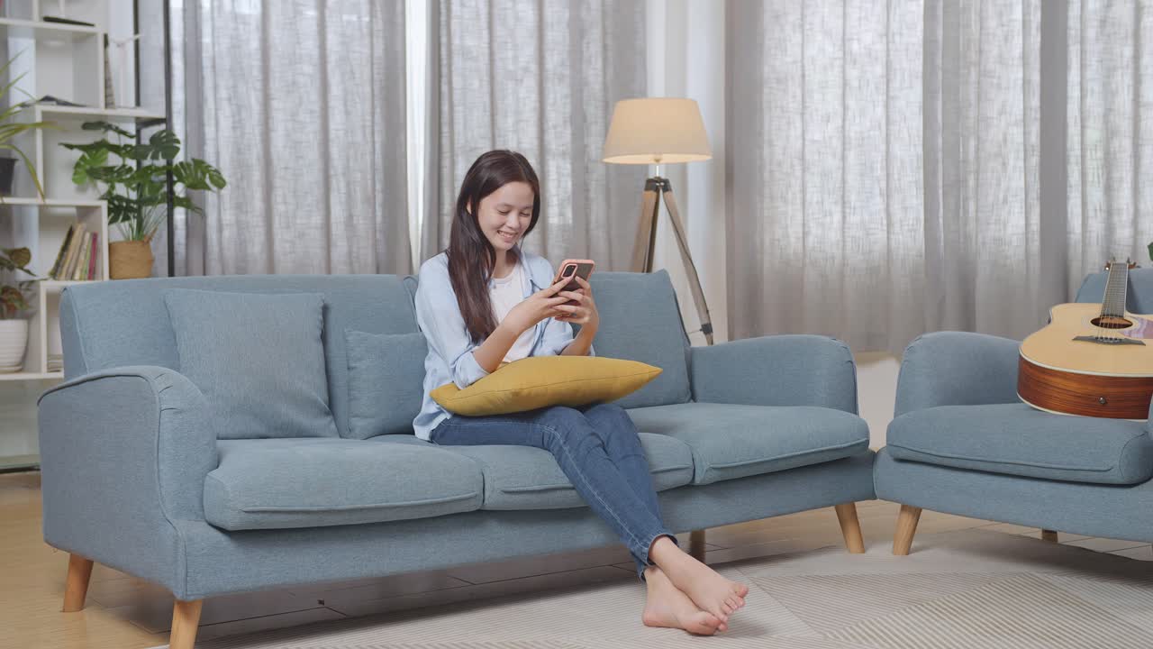 아시아 십대 소녀의 몸이 가득 차 집에서 거실에서 소파에 앉아 스마트폰을 즐기고 있습니다.