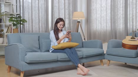 full body of asian teen girl enjoys playing smartphone while sitting on sofa in the living room at home