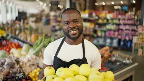 Retrato-De-Un-Hombre-De-Piel-Negra-Con-Una-Camiseta-Marrón-Y-Un-Delantal-Negro-Posando-Con-Una-Caja-De-Manzanas-De-Color-Amarillo-Verdoso-En-Las-Manos-En-Un-Supermercado