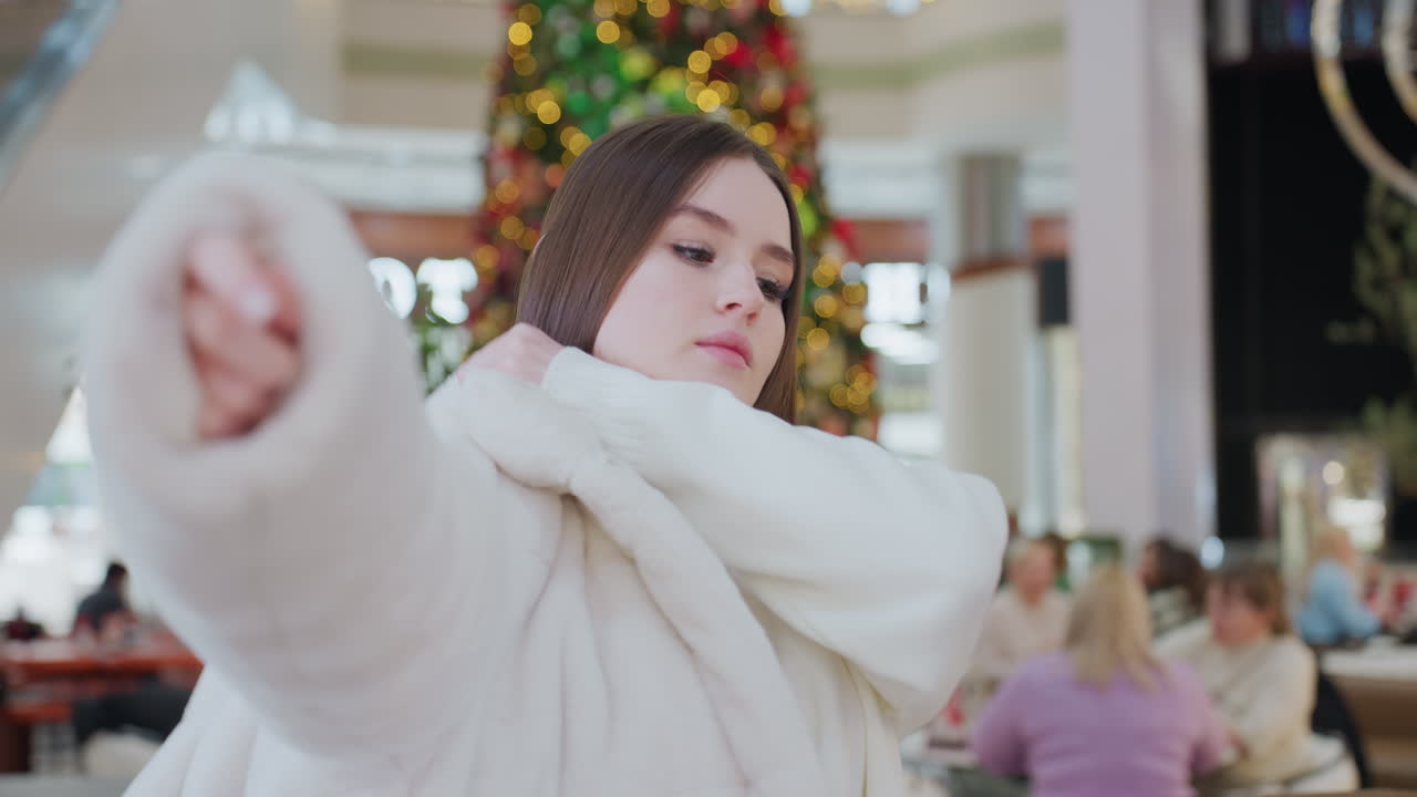 Woman putting on winter jacket in stylish shopping mall decorated with festive Christmas tree and warm lights, other shoppers are seated in the background