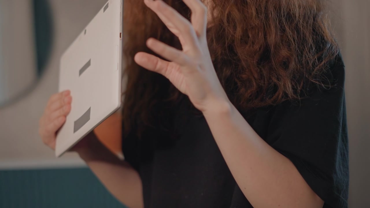 Vista cercana de una persona con cabello largo y ondulado sosteniendo una tableta en la mano izquierda mientras la mano derecha gesticula con los dedos curvados y extendidos, sugiriendo tocar, deslizar, explicar y una interacción digital activa.