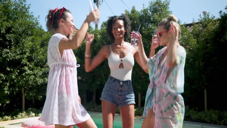 laughing female friends enjoying drinks and dancing by pool on sunny day