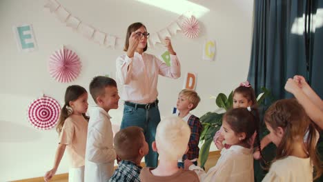 Una-Profesora-Rubia-Con-Un-Peinado-Bob,-Gafas,-Camisa-Blanca-Y-Vaqueros,-Imparte-Una-Clase-Para-Niños-En-Edad-Preescolar-En-La-Que-Explica-Cómo-Estirar-Los-Brazos-Correctamente-Y-Realizar-Un-Breve-Calentamiento-Entre-Lecciones-Para-Que-Los-Niños-No-Se-Queden-Demasiado-Tiempo.