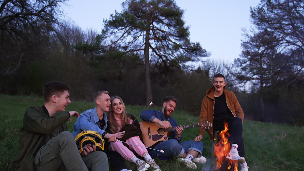 Youngsters having picnic near the bonfire in the forest. Guys are roasting marshmallows on fire, speaking and laughing.