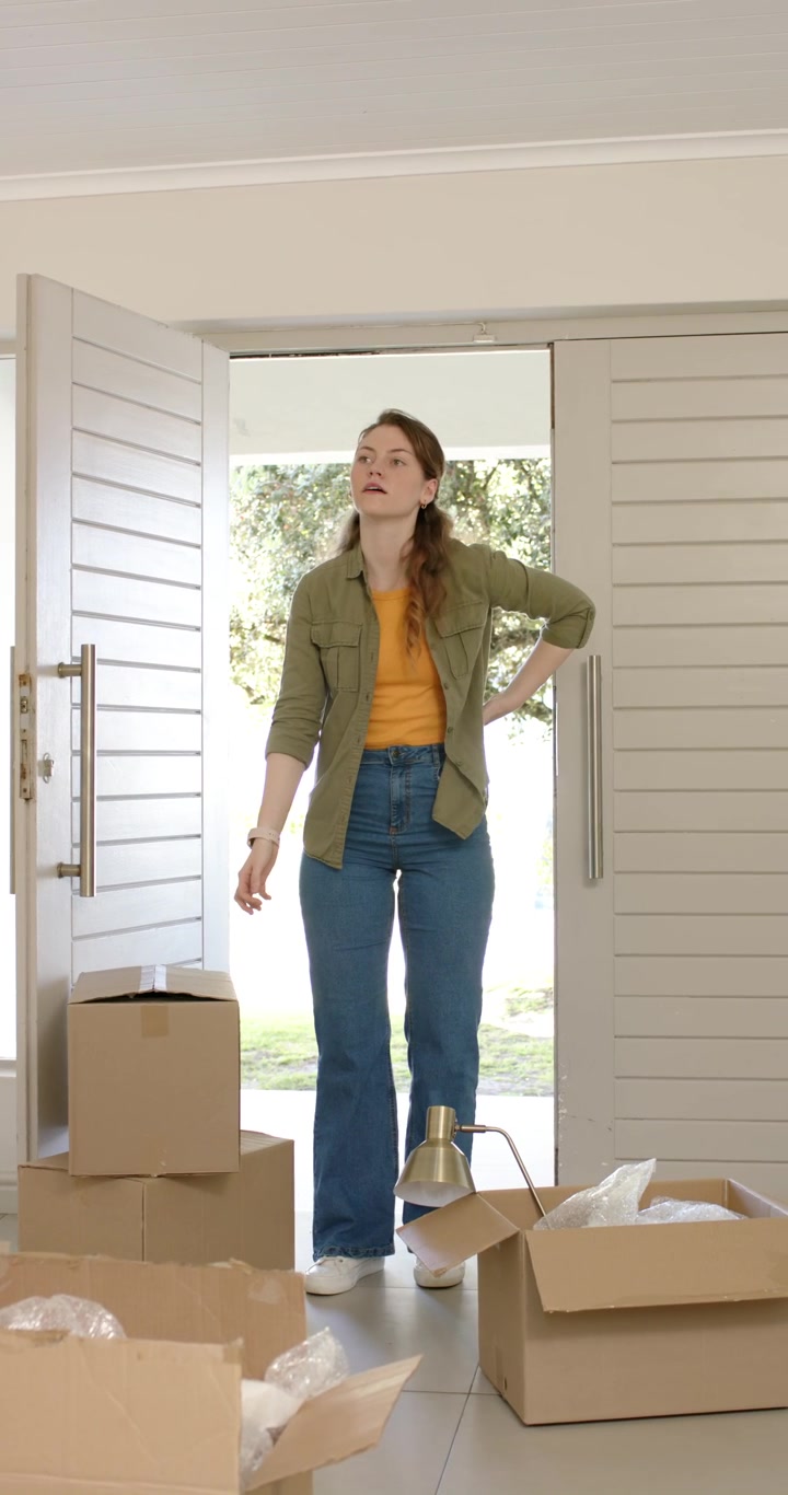 Vertical video: White woman arriving at door carrying box and stacking moving boxes surveying foyer