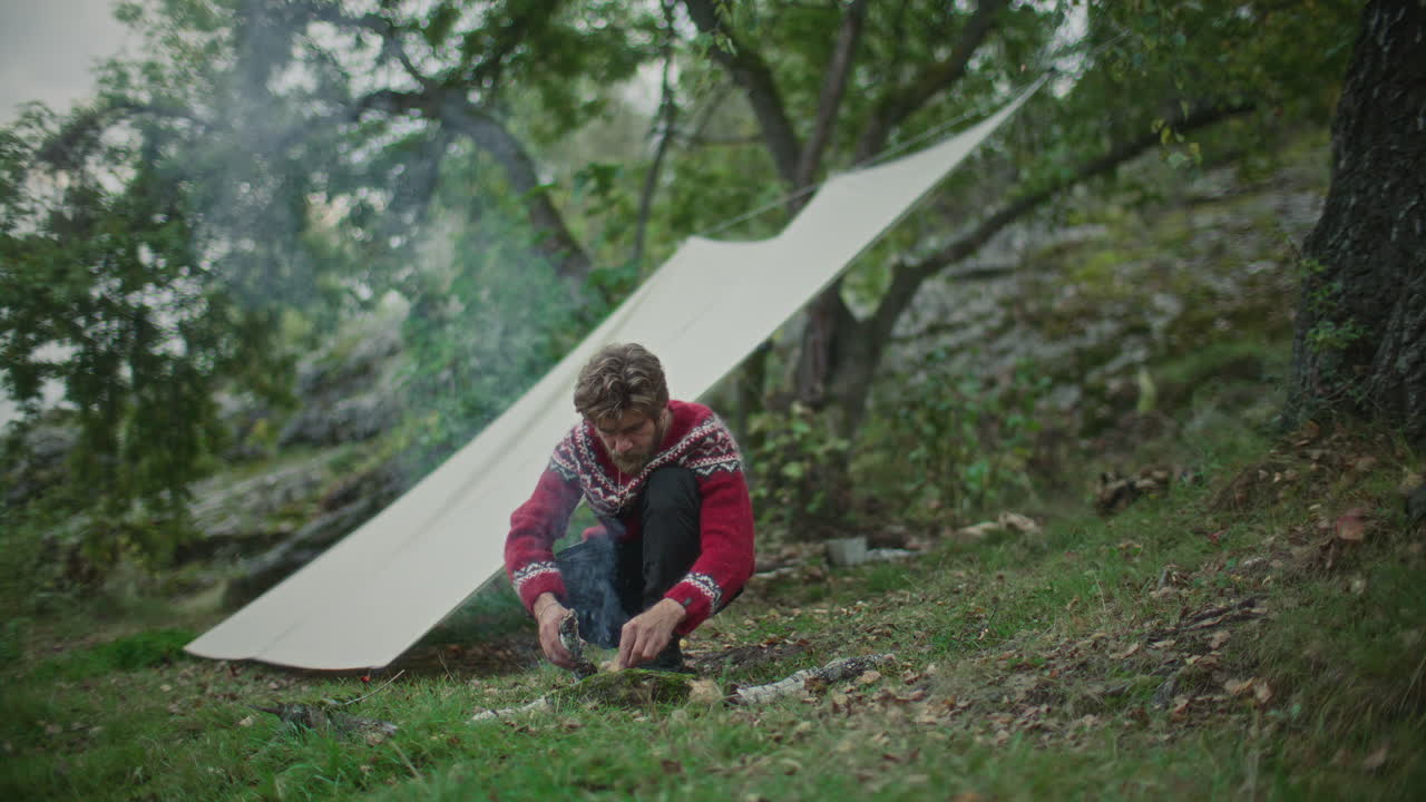 Man Making Campfire with Firewood beside Tarp Shelter in Forest