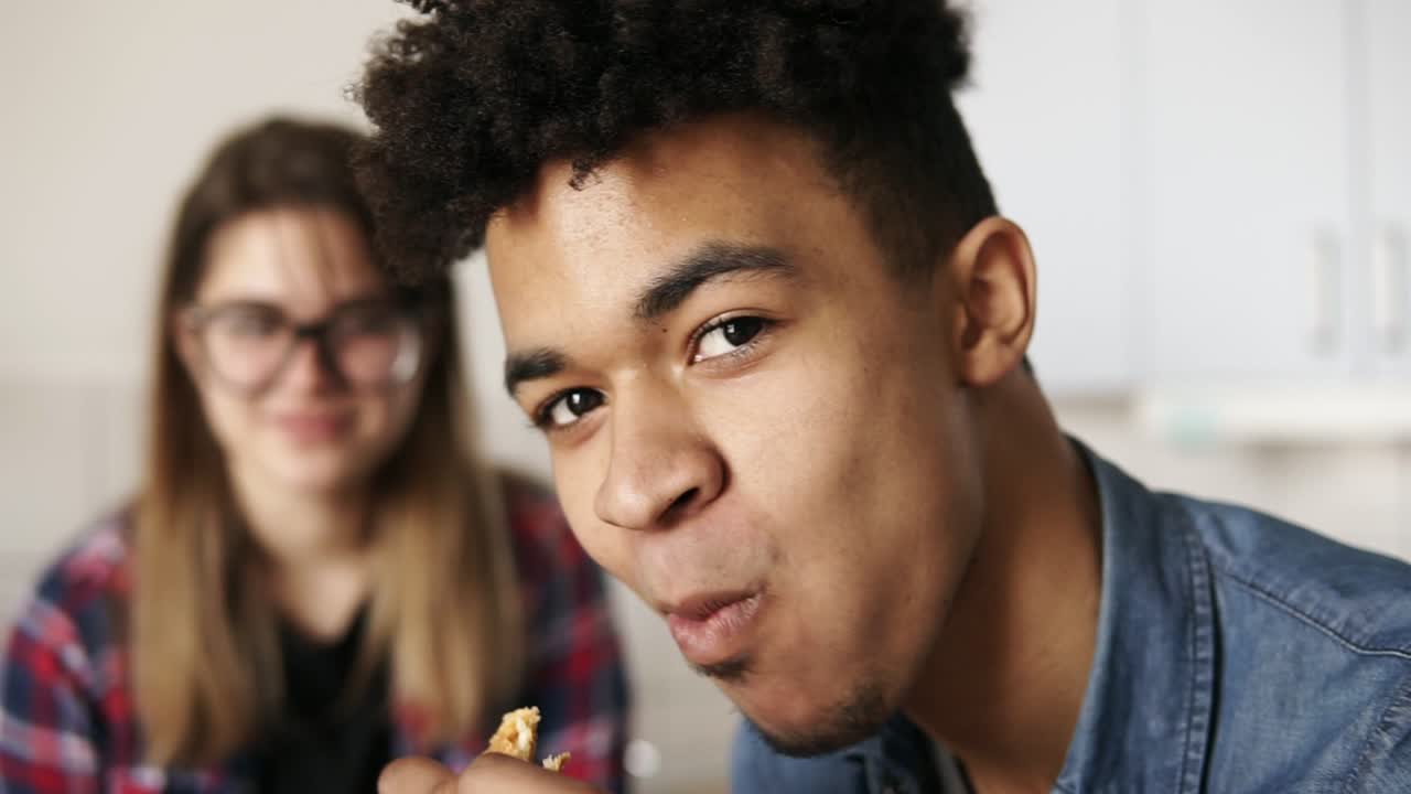 Close up footage of a handsome mulatto guy in his 20's with expressive deep dark brown eyes looking at the camera, eating croissant, with his girlfriend sitting in the background.