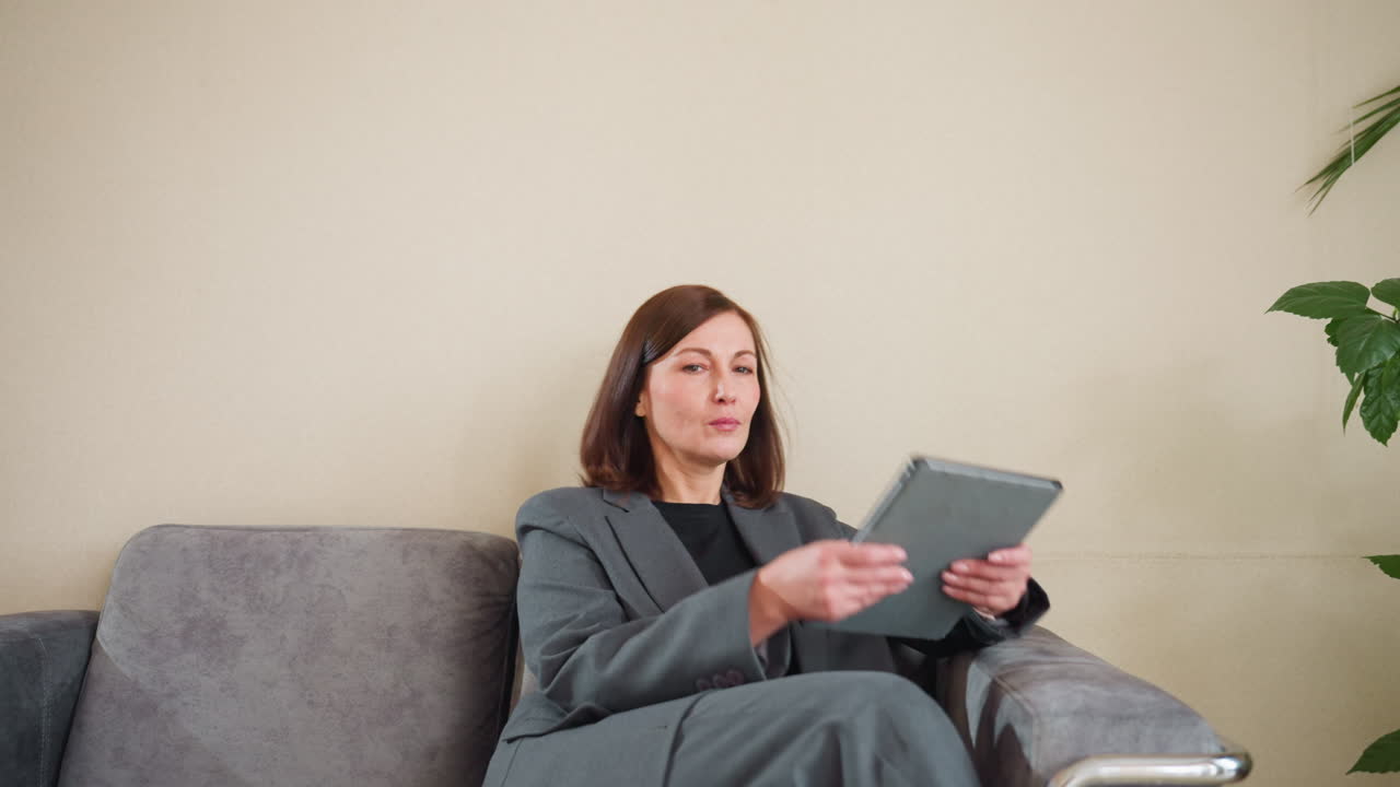 Side profile of businesswoman with brown hair wearing gray blazer looking forward in office setting. Professional female standing beside potted plants and palm decor against cream wall background