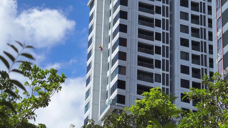 Vista lejana de un trabajador de acceso por cuerda pintando o manteniendo el exterior de un edificio masivo de gran altura, enfatizando la altura extrema, el riesgo laboral y la escala dramática de la torre urbana
