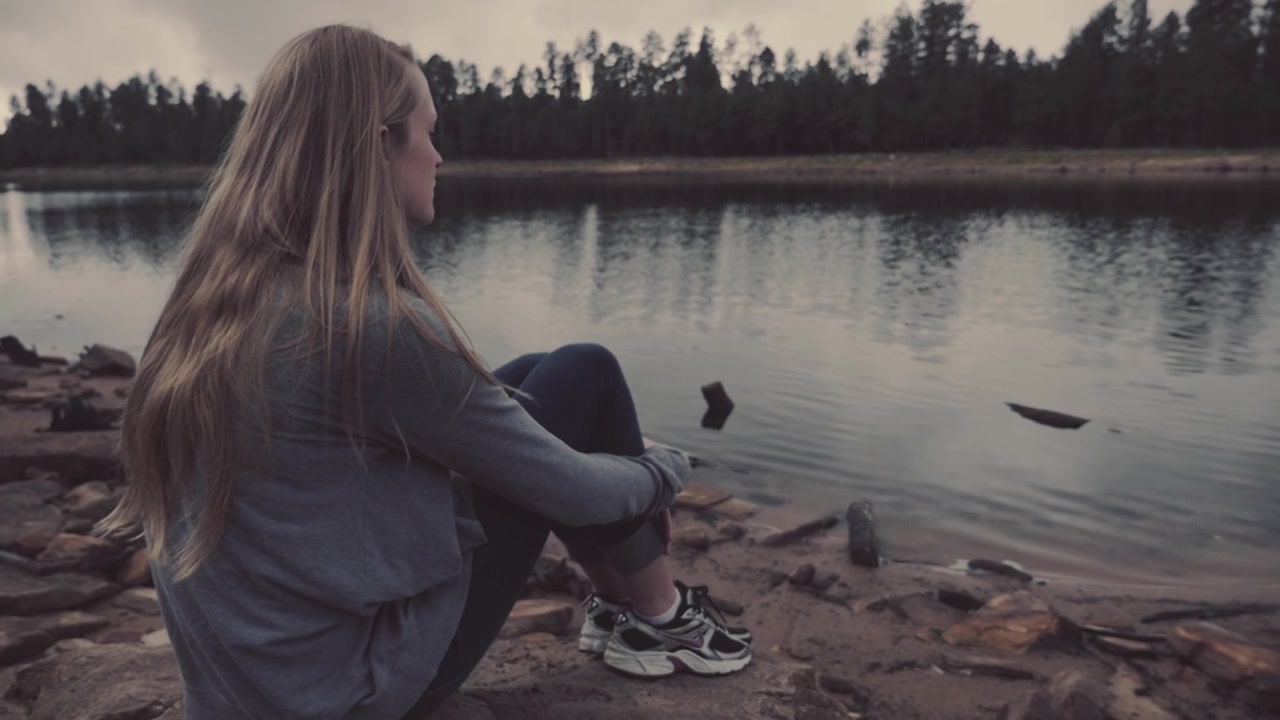 Young Woman Sits Peacefully by a Lake Shore on a Cloudy Day