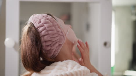 woman in bandage relaxing with cosmetics in bathroom. brunette female with hair in ponytail makes face tightening mask in bathroom slow motion