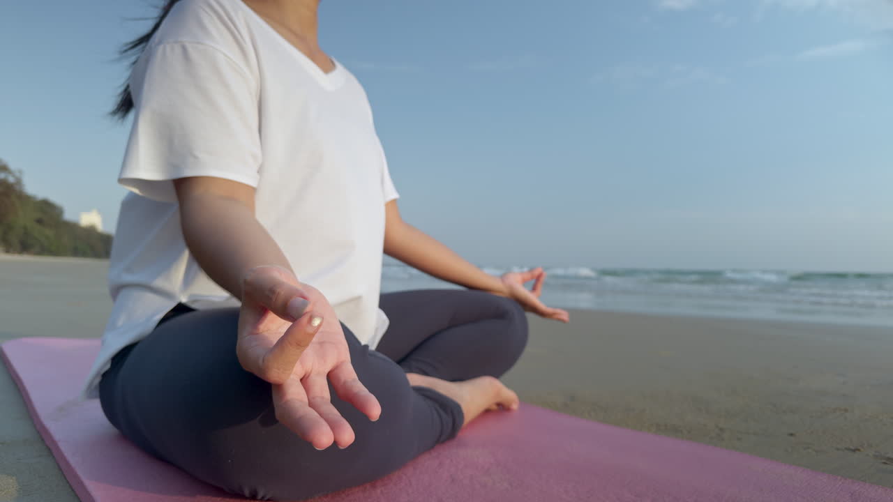 Detailní záběr ženy praktikující jóga meditaci na pláži při východu slunce. Zaměřeno na ruce v mudra gestu, s klidným oceánem v pozadí, koncept všímavosti, relaxace a duševního zdraví.