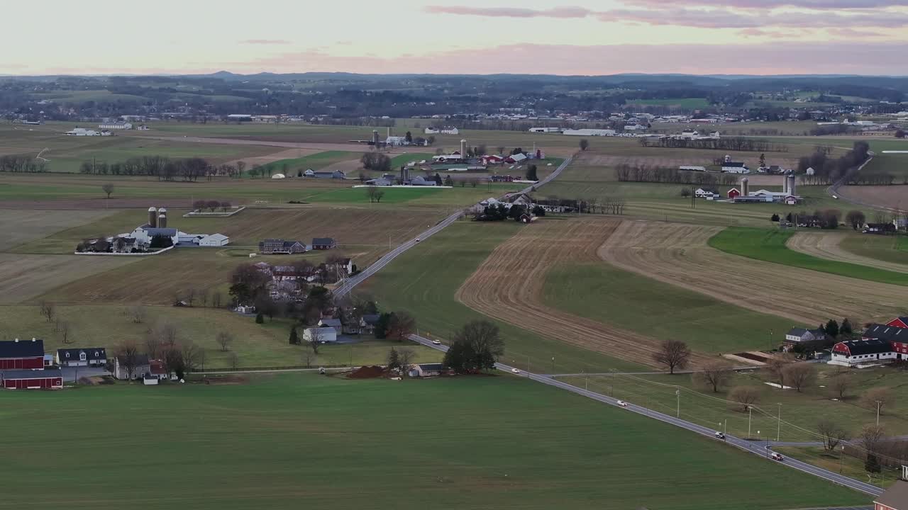 Traffic on intersection road in rural area of North Carolina with colorful agricultural fields in winter. Farms with silo and stable in USA. Aerial wide shot. Sunset time.
