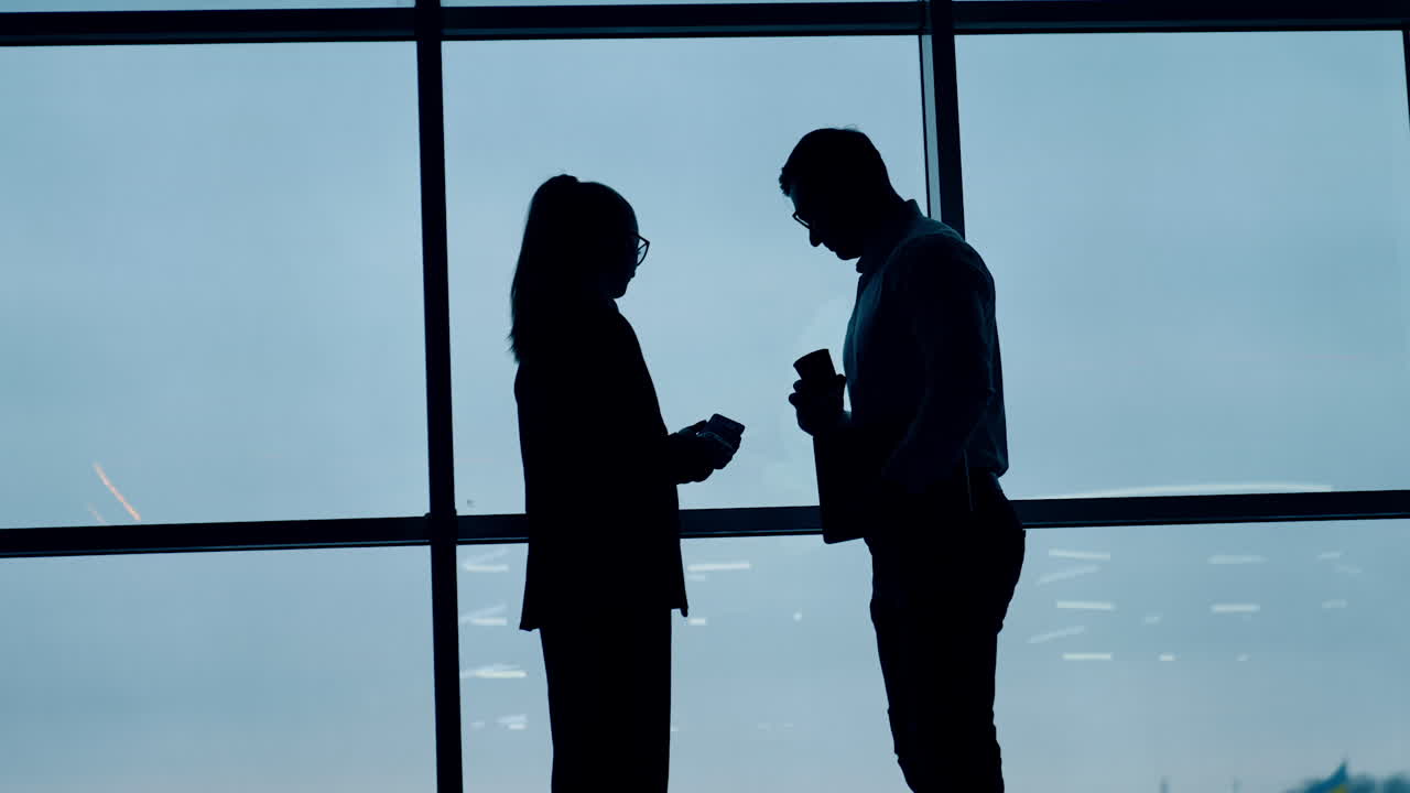 Colleagues' silhouettes in the office. Man with coffee cup looks at the phone in woman's hand. Panoramic window at backdrop.