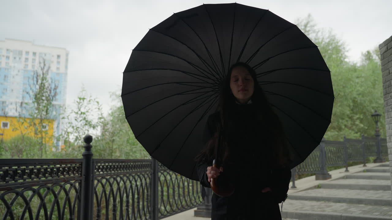 Donna con ombrello nero che cammina lungo il camminamento sul fiume, oltrepassando ringhiere decorate e lampioni sotto un cielo nuvoloso con gocce di pioggia, atmosfera contemplativa, cappotto elegante e grande parasole in controluce