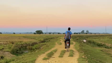 young village boy with backpack cycle in rural path through agriculture fields in bangladesh, back view