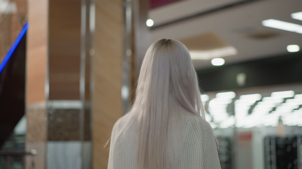 back view of blonde shopper strolling through modern mall hall wearing knitted white sweater straight hair cascading down under bright lights as man approaches moving walkway in background