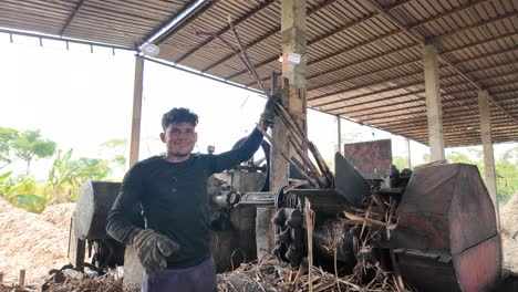 man working on a sugarcane mill, holding sticks with a smile in a rural setting