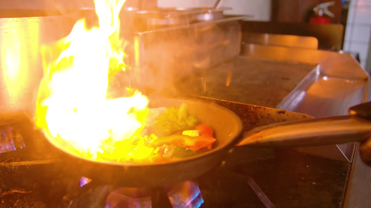 Chef che aggiunge verdure fresche e verdi in padella, con grandi fiamme che divampano