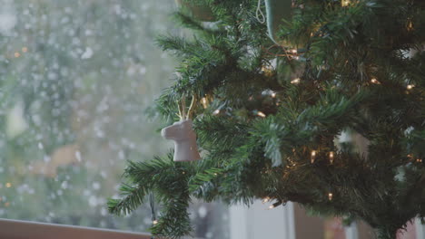 christmas tree decorated in white with lights stands near window while it snows