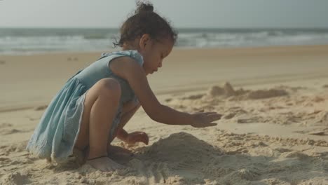 The-girl-sits-on-the-sandy-shore-and-builds-a-sand-castle---Happy-little-girl-plays-with-sand-on-the-beach-in-the-background-at-the-blue-sea-at-sunset