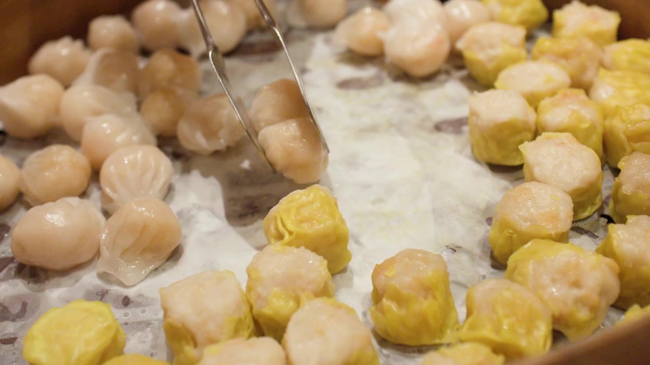 Foto close-up penjepit logam mengambil pangsit udang dan shumai dari kukusan bambu besar