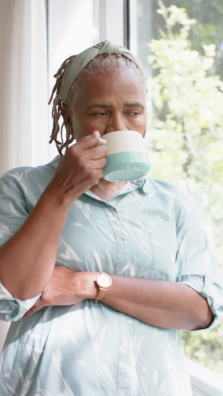 Video verticale di una donna anziana afroamericana pensierosa con una tazza di caffè a casa, al rallentatore.