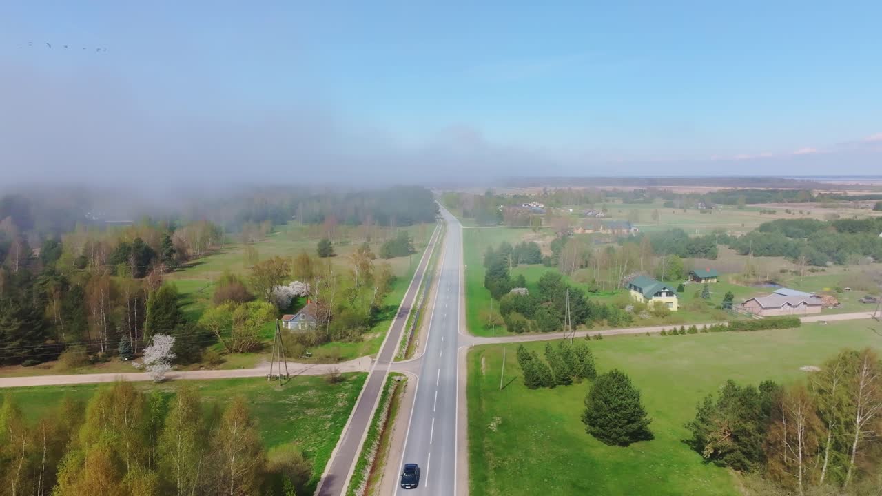 Low fog creeps over fields near Liepāja as drone pulls back along rural highway