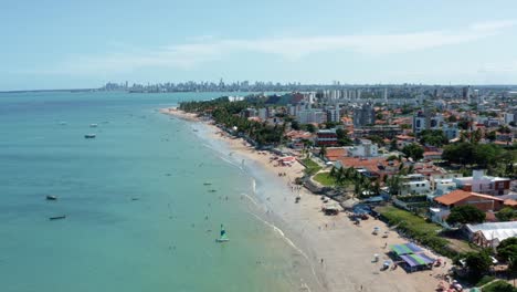 dolly in tilting up aerial extreme wide shot of the tropical bessa beach in the capital city of joao pessoa, paraiba, brazil with people enjoying the ocean surrounded by palm trees and buildings