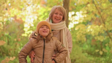 mother shares tender moment with daughter in sunlit forest, smiling warmly while gently holding child close, golden autumn foliage glowing in background