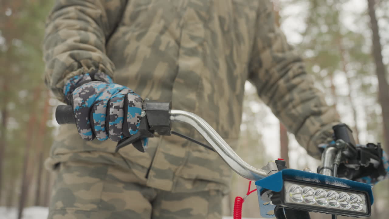 Gloved hand gripping atv handlebar close, blue camo glove on throttle, brake lever and LED bar illuminated, camo jacket and helmeted rider in snowy pine backdrop, tense preparation for extreme ride