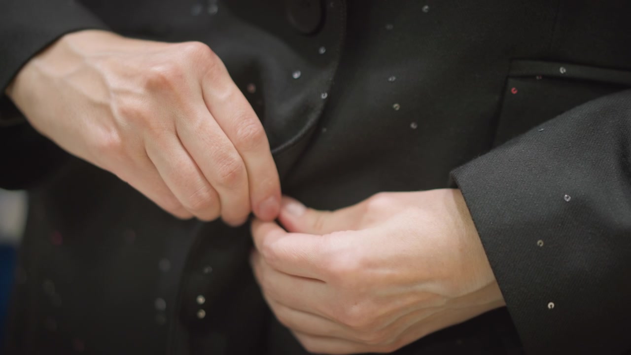 Close up of hands fastening black jacket with small reflective dots. person manipulates fabric and buttons demonstrating precise manual dexterity against soft lighting. Background remains
