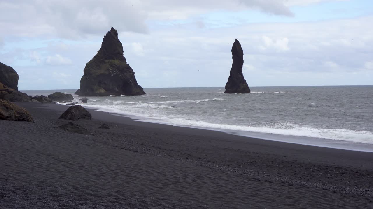 playa de arena negra con altas pilas de mar bajo un cielo nublado en islandia, olas golpeando la costa