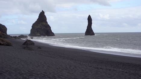 Schwarzer-Sandstrand-Mit-Hoch-Aufragenden-Felsnadeln-Unter-Einem-Bewölkten-Himmel-In-Island,-Wellen-Plätschern-Ans-Ufer