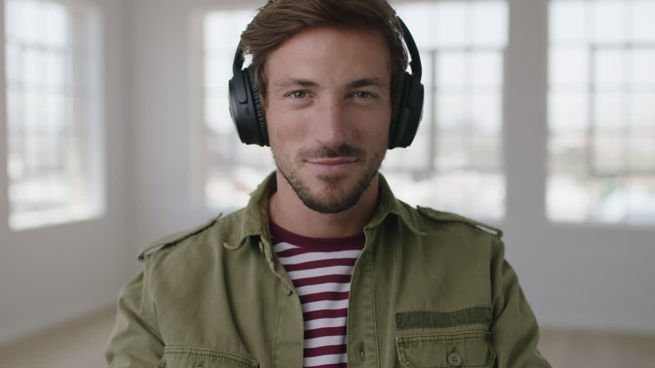 close up portrait of attractive young man smiles at camera puts on headphones listeningto music enjoying leisure activity in apartment
