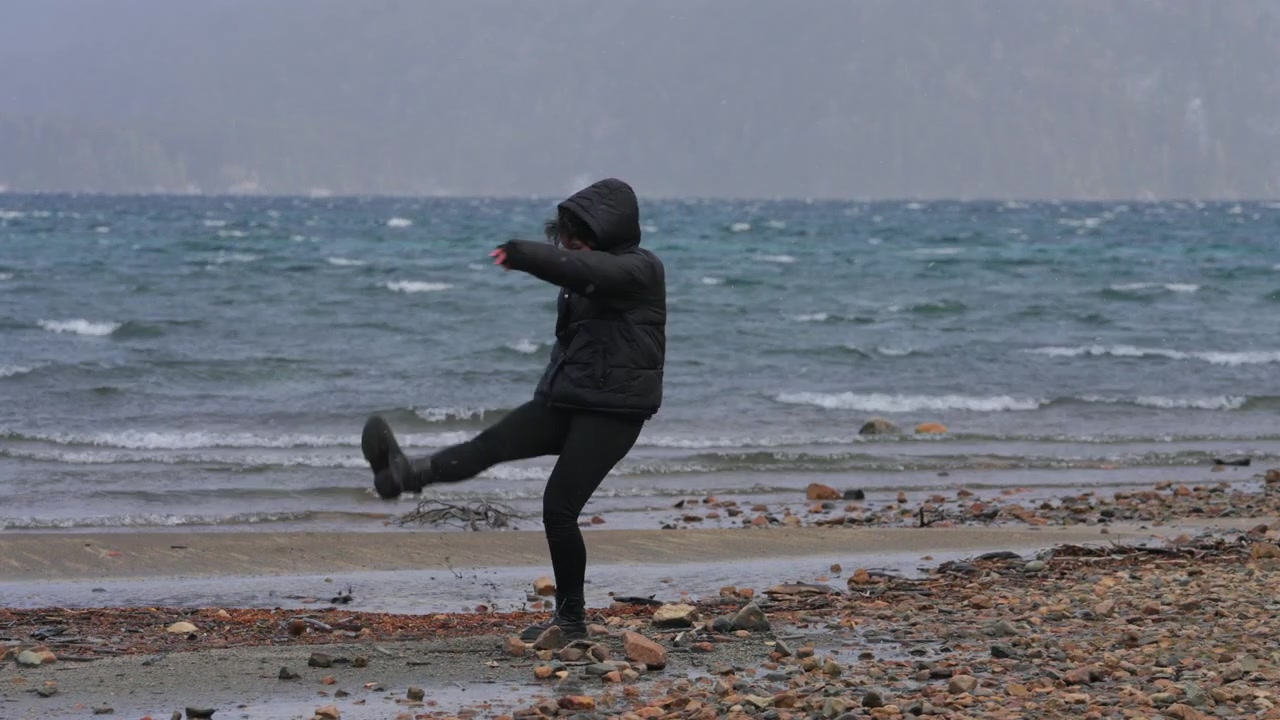 Ceketine sarınmış bir kadın, arka planda dalgaların olduğu kayalık bir plajda yürüyor. Sahne, soğuk ve rüzgarlı bir günü yansıtıyor. Yalnızlık veya kış seyahati temaları için uygun.