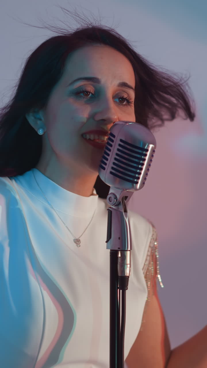 A singer in a white dress passionately sings, hands in motion, in front of a vintage microphone against a white background. Her expressive face and flowing hair add to the emotional