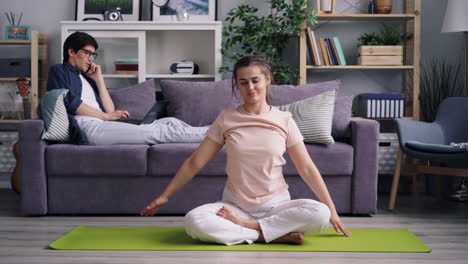 woman practicing yoga at home while partner relaxes on the couch.