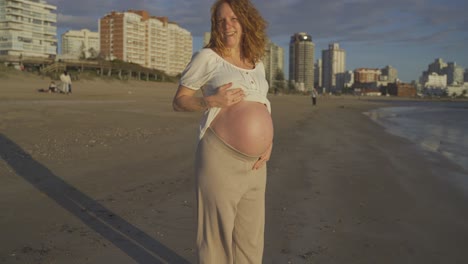 mujer pelirroja caucásica embarazada que muestra una gran barriga mientras camina en la playa al atardecer, punta del este uruguay